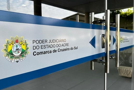 Foto da porta de entrada do Fórum de Cruzeiro do Sul. É uma porta de vidro com adesivo em tons azuis com logomarca do Estado e escrito "Poder Judiciário do Estado do Acre: Cruzeiro do Sul".