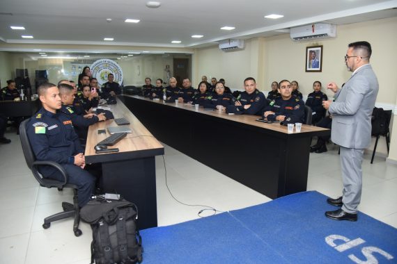 A imagem mostra uma palestra ou treinamento sendo ministrado em uma sala de situação e gerenciamento, voltada possivelmente a agentes de segurança pública. Descrição da cena: Um homem de terno cinza claro, óculos e gravata está em pé, gesticulando enquanto fala para um grupo de pessoas uniformizadas, indicando que é o palestrante ou facilitador da atividade. Os participantes estão sentados ao redor de uma mesa longa e retangular, usando uniformes escuros com insígnias e bandeiras do Brasil nos braços, sugerindo que são membros de alguma corporação — provavelmente policiais militares ou bombeiros. Sobre a mesa há copos descartáveis, garrafas de água e bonés do uniforme, indicando que é uma sessão relativamente longa e informal. Ao fundo, uma grande tela exibe uma apresentação com os termos “Mundo VUCA” e “Mundo BANI”, conceitos usados para descrever contextos modernos de instabilidade, incerteza e complexidade, bastante aplicados em ambientes de liderança e estratégia. Parte da frase visível na tela diz: “Como podemos definir ou entender nossa realidade atual?”, o que sugere que o tema da palestra está relacionado a cenários contemporâneos, tomada de decisão e adaptação em tempos de mudança. Ambiente: A sala possui acabamento moderno, com painéis de madeira e ar-condicionado. O letreiro ao fundo indica que é a “Sala de Situação e Gerenciamento”, reforçando o caráter tático e estratégico da reunião.